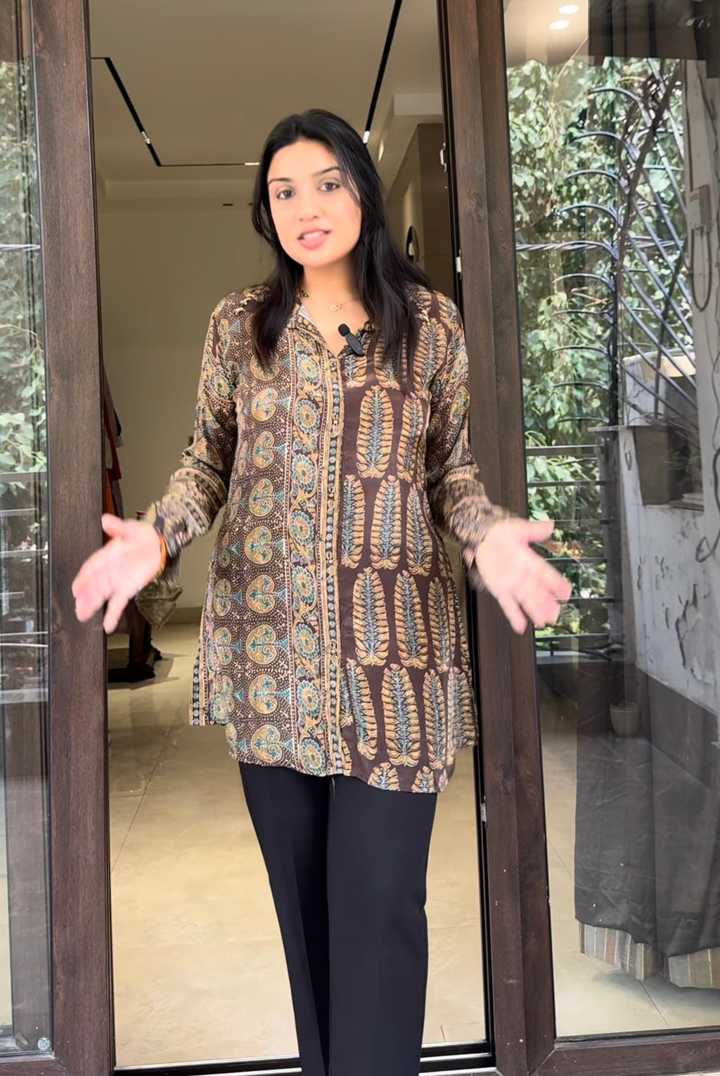 Woman wearing a patterned shirt and black pants standing in front of a glass door.